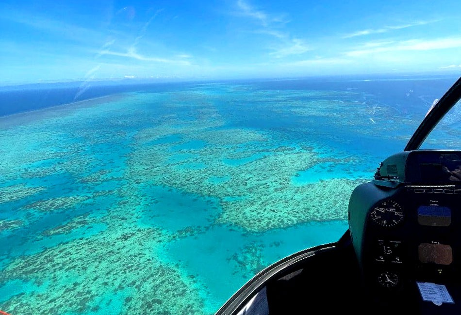 Aerial view from a helicopter cockpit showing clear blue ocean water and coral reefs below under a sunny sky.