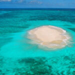 A small, sandy island sits in the middle of clear turquoise ocean water under a blue sky.