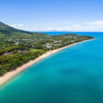 Aerial view of a coastline with turquoise water, sandy beach, green forest, buildings, and a mountain under a clear blue sky.