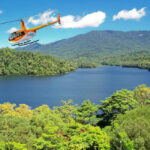 An orange helicopter flies over a lush green forest and a large lake, with mountains and a blue sky in the background.