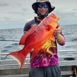 Person standing on a boat holding a large, bright orange-red fish with the ocean and cloudy sky in the background.