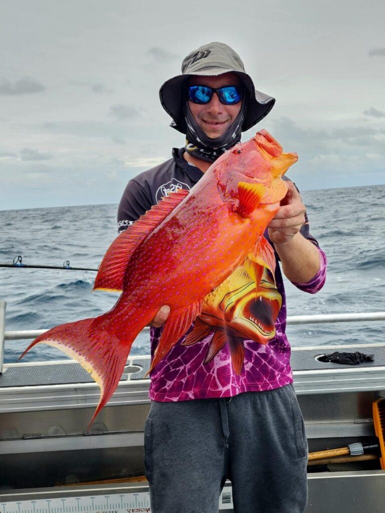 Person standing on a boat holding a large, bright orange-red fish with the ocean and cloudy sky in the background.