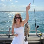 A woman in a white dress stands on a boat holding up a freshly caught fish with one hand; the ocean and distant shoreline are visible in the background.