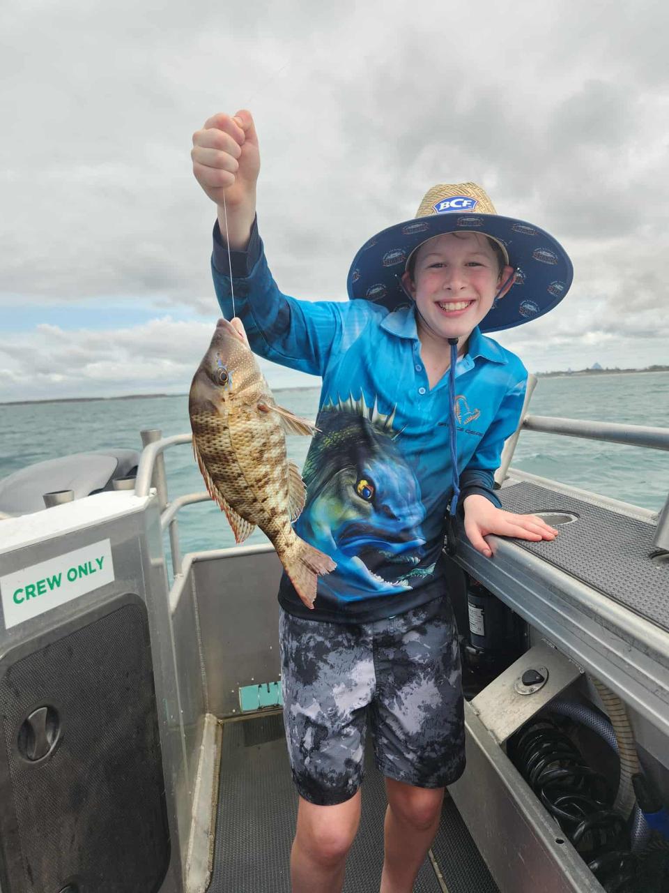 A smiling child on a boat holds up a freshly caught fish with one hand, wearing a blue fishing shirt, shorts, and a wide-brimmed hat.