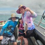 Three people on a boat, one adult holding a small fish on a fishing line, two children stand beside her, all smiling, wearing hats and colorful clothing. Ocean and cloudy sky in background.