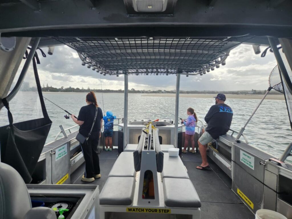 Four people, including two children, are fishing from a boat on calm water under a cloudy sky; safety and warning signs are visible inside the boat.