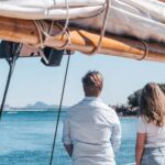 Two people stand on a boat, facing the water and distant shoreline, with sail rigging overhead and clear blue skies.