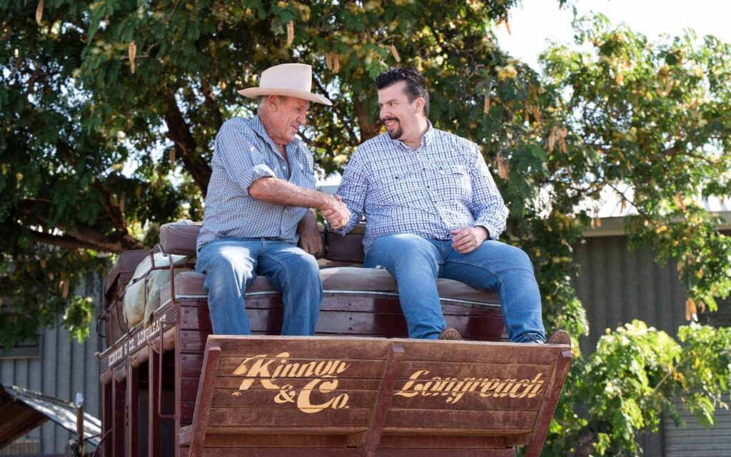 Two men in casual attire shake hands while sitting on a wooden wagon labeled "Kinnon & Co. Longreach," with trees and a building in the background.