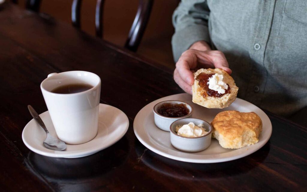 A person spreads cream and jam on a biscuit, with a cup of tea, extra jam, and cream on a plate on a wooden table.