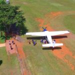 A small plane is parked on a grassy field as several people walk from the aircraft toward a nearby fence and trees.