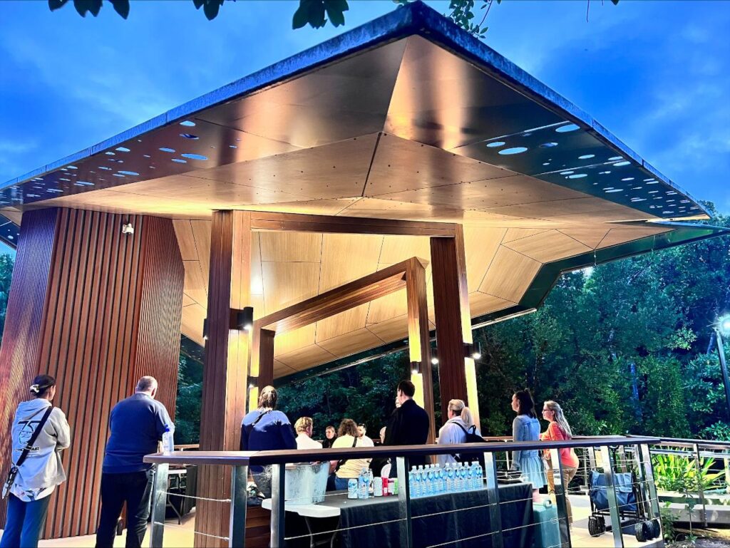 A group of people stands under a modern, angular pavilion with a wooden roof, gathered around tables with bottled water in an outdoor setting at dusk.