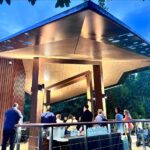 A group of people stands under a modern, angular pavilion with a wooden roof, gathered around tables with bottled water in an outdoor setting at dusk.
