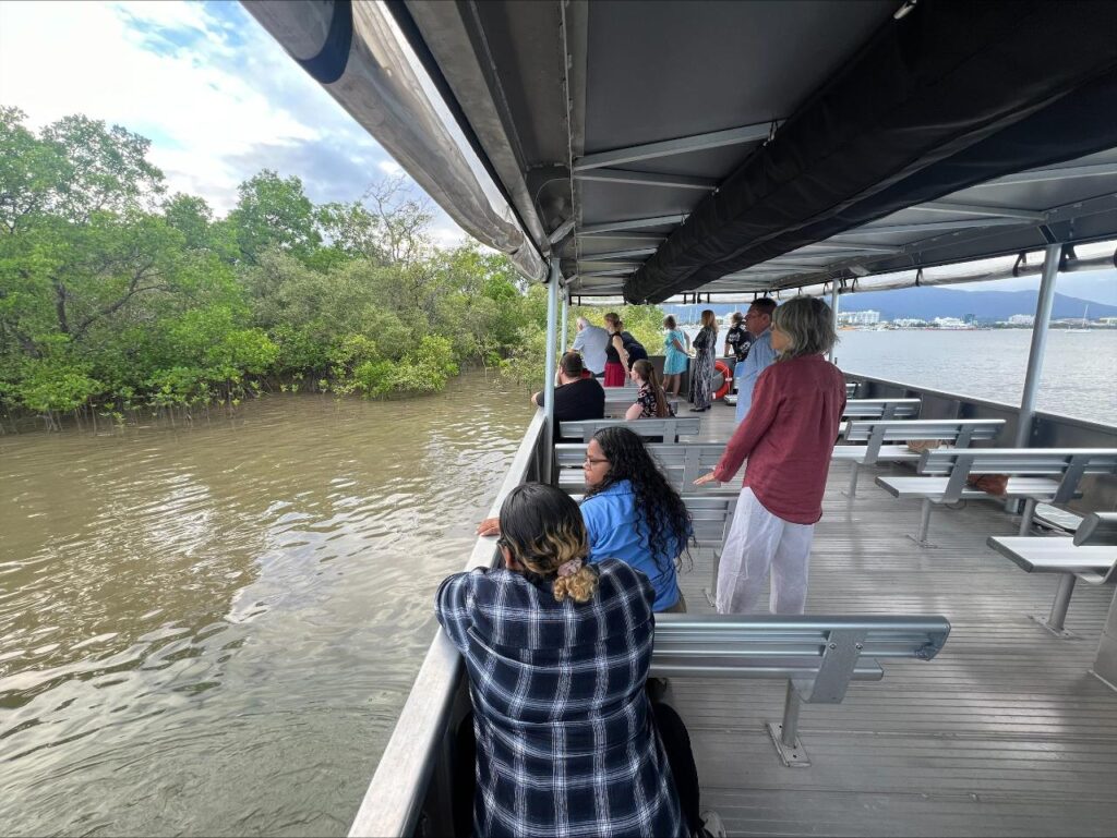 A group of people stand and sit on a covered boat deck, looking out at water and green mangroves along the shore.