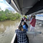 A group of people stand and sit on a covered boat deck, looking out at water and green mangroves along the shore.