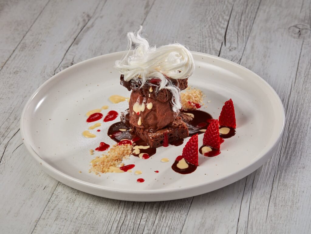 A plated dessert featuring chocolate ice cream, red sauce, fresh strawberries, white sauce drizzle, crumbled nuts, chocolate pieces, and a garnish of spun sugar on a white plate.