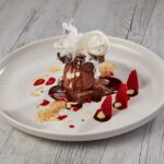 A plated dessert featuring chocolate ice cream, red sauce, fresh strawberries, white sauce drizzle, crumbled nuts, chocolate pieces, and a garnish of spun sugar on a white plate.