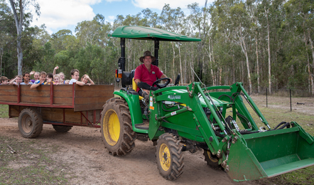A man drives a green tractor pulling a wooden trailer filled with children through a rural, wooded area on a dirt path.