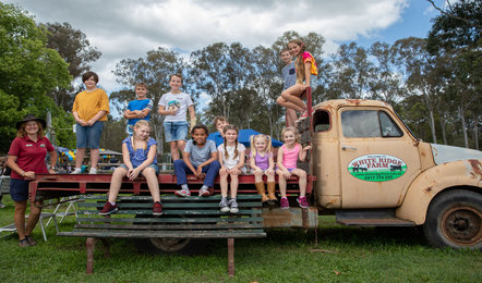 A group of children and one adult sit and stand on an old flatbed truck labeled “Water Ridge Farm” in an outdoor setting with trees and grass.