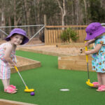 Two young children wearing hats and summer clothes play mini-golf on a green outdoor course surrounded by wood and gravel, with trees in the background.