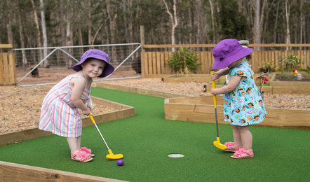 Two young children wearing hats and summer clothes play mini-golf on a green outdoor course surrounded by wood and gravel, with trees in the background.