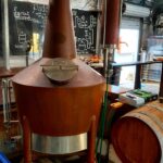 Large copper brewing kettle with a round hatch on top, situated in a brewery with a wooden barrel and chalkboard menu in the background.