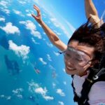 A person wearing skydiving gear smiles while free-falling above a blue ocean with scattered clouds visible below.