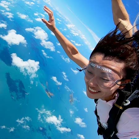 A person wearing skydiving gear smiles while free-falling above a blue ocean with scattered clouds visible below.