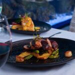 A plated gourmet meal with roasted meat, potatoes, and vegetables on a black dish, accompanied by glasses of red and white wine on a white tablecloth.