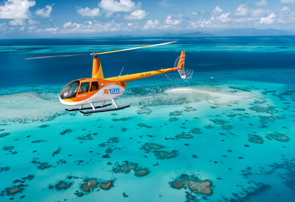 A yellow and orange helicopter flies over clear turquoise water and coral reefs near a small sandbar under a partly cloudy sky.