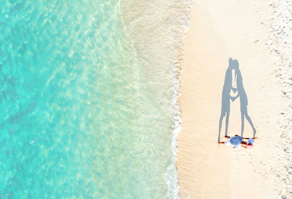 Aerial view of two people lying on a sandy beach near clear turquoise water, with their shadows forming the shape of a heart.