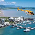 A yellow and orange helicopter flies over a coastal city marina with docks, boats, and nearby buildings; forested hills are visible in the background.