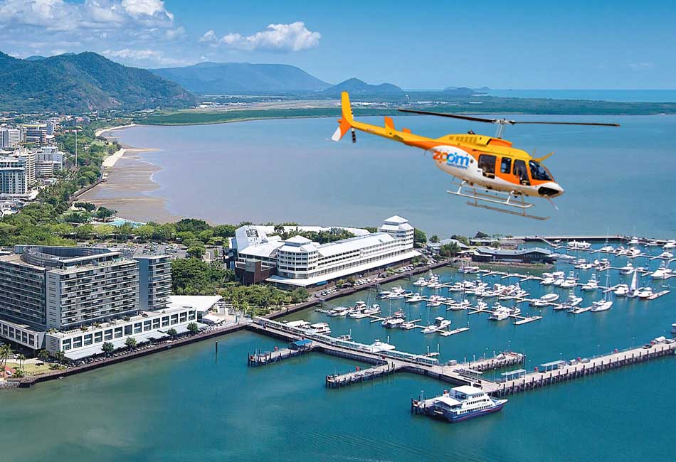 A yellow and orange helicopter flies over a coastal city marina with docks, boats, and nearby buildings; forested hills are visible in the background.